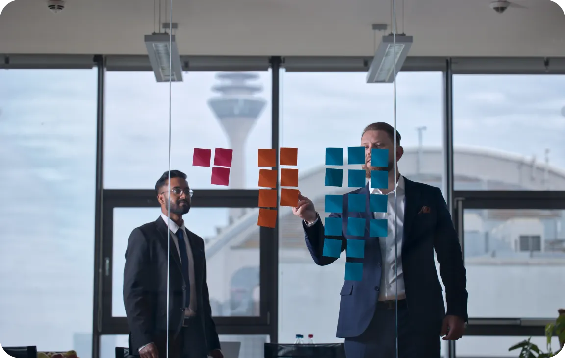 Two professionals in a strategy session with colorful sticky notes on glass, Düsseldorf TV tower visible outside.
