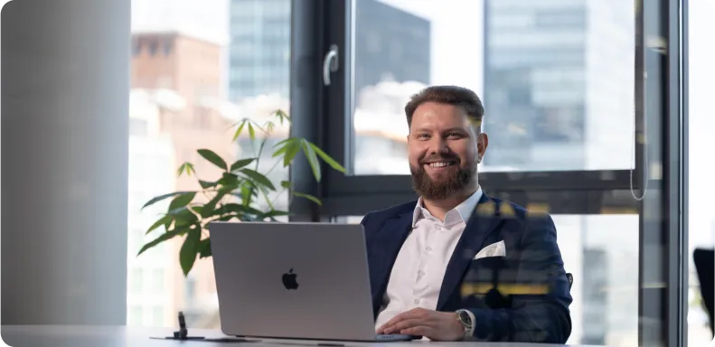 A smiling bearded man in a business suit works on a MacBook at a desk with a potted plant nearby, in an office with a view of city buildings through the window.